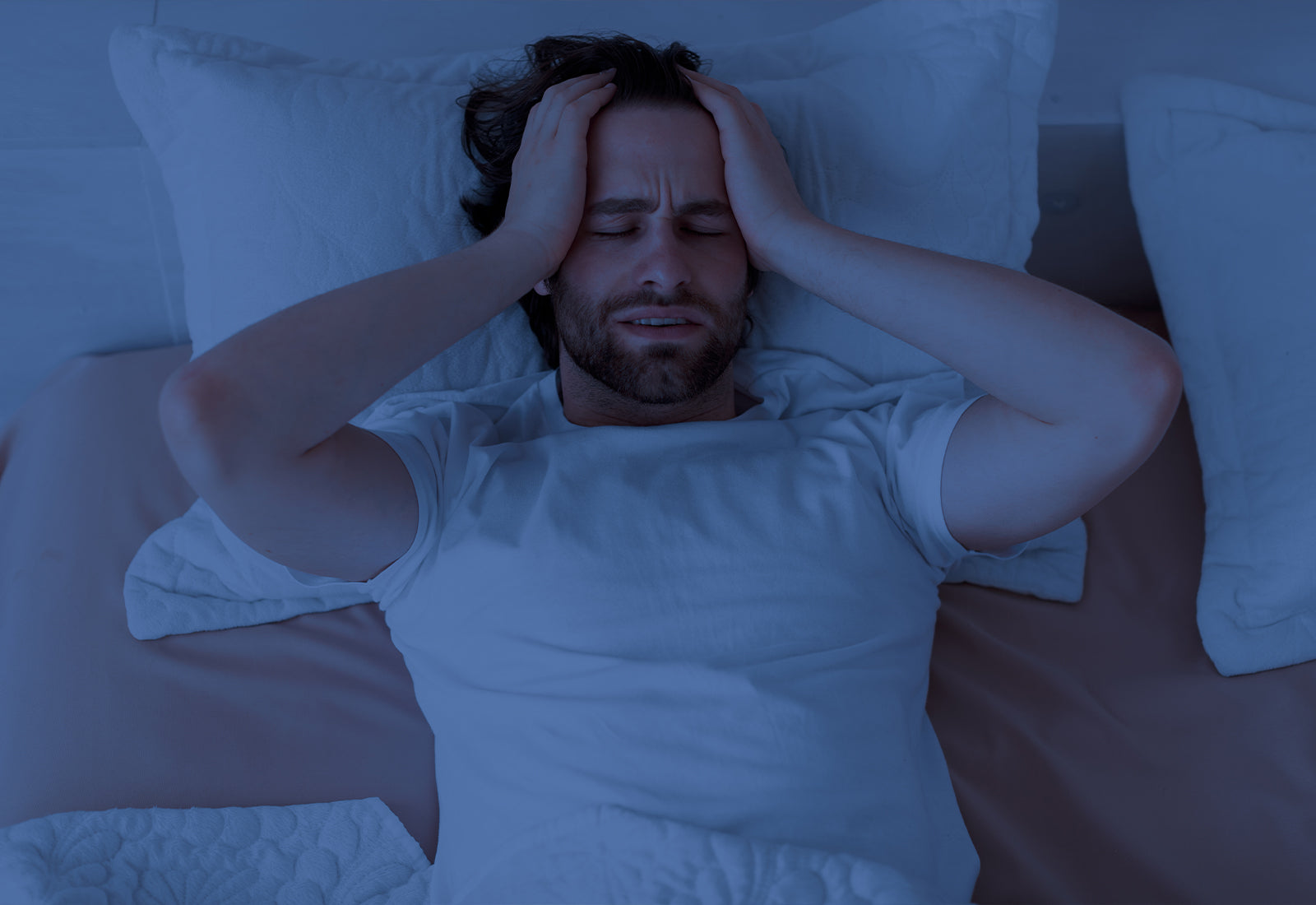 A man lying in bed at night, holding his head with both hands, appearing to have trouble sleeping